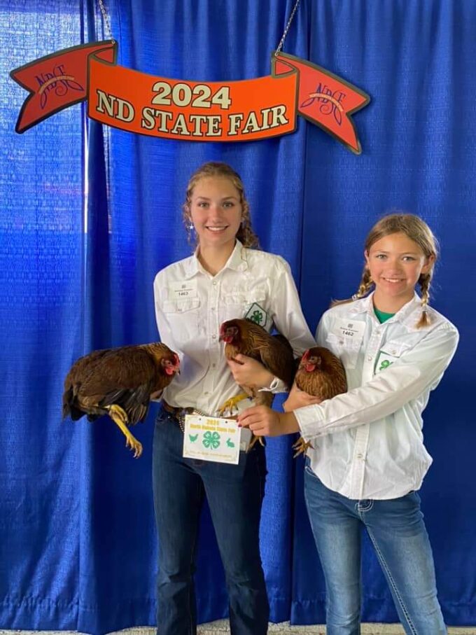 Two girls hold Welsummer Chickens at the 2024 ND State Fair, standing in front of a blue curtain and event sign.