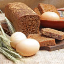 A loaf of brown bread, slices of bread, three eggs, and wheat stalks on a rustic surface.