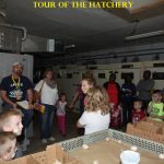 A group of adults and children listen to a guide inside a hatchery room with eggs and equipment visible.