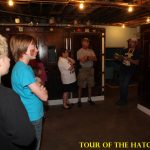A group of people listens to a guide inside a dimly lit hatchery building. Yellow text reads "TOUR OF THE HATCHERY.