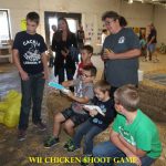 Several children and an adult watch as two boys play a Wii Chicken Shoot game in a barn-like indoor setting.