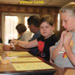 Children and adults sit at a table playing a game with yellow cards and small containers of markers. "WINGO GAME" is written above.