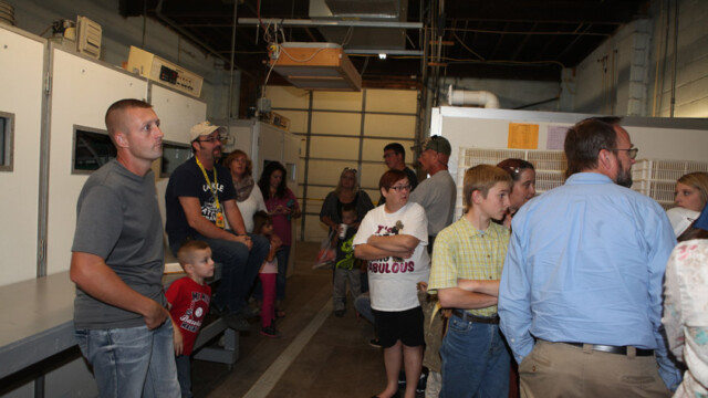 A group of people stand and sit in an industrial room, listening to someone outside the frame.