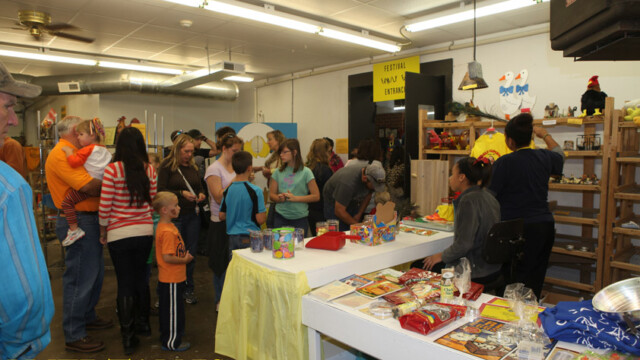 People line up inside a room at a prize table with various items, toys, and cans displayed on tables and shelves.