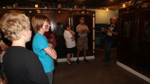A group of people listens to a guide inside a dimly lit hatchery building. Yellow text reads "TOUR OF THE HATCHERY.