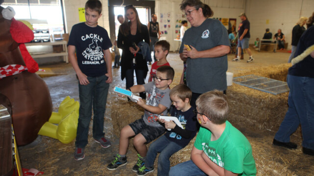 Several children and an adult watch as two boys play a Wii Chicken Shoot game in a barn-like indoor setting.
