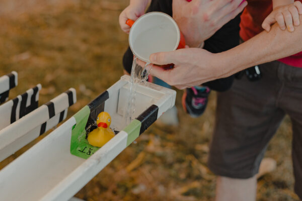 A person pours water into a trough to move a toy rubber duck in a race game while holding a child.