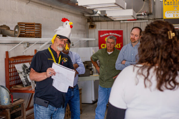 A man wearing a chicken hat holds papers and speaks to a group of four people in an indoor setting.