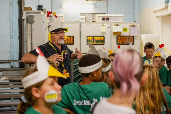 An adult speaks to children wearing paper chicken hats in a room with machinery and equipment in the background.